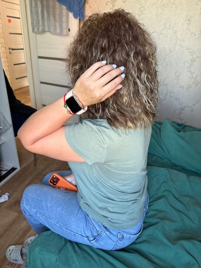 In this media file, a person is captured from a high-angle perspective, lying down with their head resting on their hand. The person has curly, brown hair with lighter streaks and light blue nail polish. They are wearing a light green t-shirt and blue jeans. A pink smartwatch is visible on their left wrist, adorned with a red string bracelet and beads. An orange smartphone is held in their right hand. The background consists of a light-colored wallpaper with a floral pattern and a crumpled green blanket. Furniture, possibly a dresser with drawers, is partially visible in the lower left and bottom of the frame. The scene appears to be indoors, with no specific time of day or weather cues. The person's posture and hand on their head might suggest a moment of rest or contemplation. No visible text is present in the image.