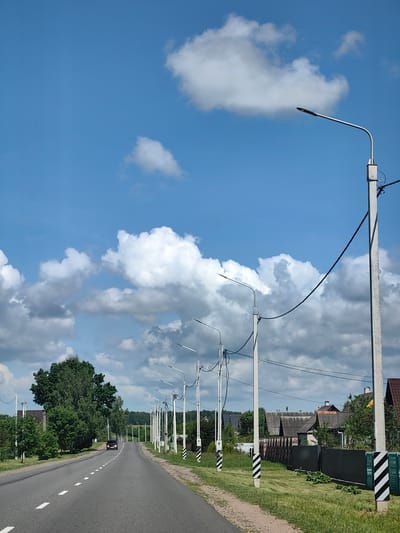 The media file depicts a rural road scene on a bright, partially cloudy day. The road stretches into the distance, lined with numerous streetlights and power poles, suggesting a somewhat developed rural area. On the right side of the road, houses with dark roofs and fences are visible. Lush green trees and grass flank the road, indicating a warm season. The sky is a vibrant blue, dotted with fluffy white clouds. A single car is driving away from the viewer down the road. The scene appears peaceful and tranquil, with no people visible. The time of day is likely daytime, given the bright sunlight and clear sky. The location is identified as Ilyanskiy selskiy Sovet, Belarus.