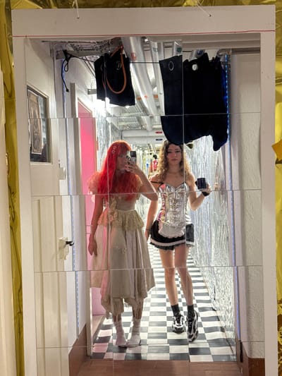 The image displays two young women reflected in a large, gridded mirror installation, located in Wallisellen, Switzerland. The mirrored surface fragments the scene into multiple squares, creating a mosaic-like view of the subjects and their surroundings.
On the upper right side of the reflection, a woman with long, wavy brown hair is visible. She wears a silver, corset-style top with black accents, black shorts or a short skirt with white trim, and black and white sneakers. She holds a smartphone up, appearing to take a selfie or record a video. Below her, a second woman with striking, long red hair is dressed in a light-colored, possibly cream or white, dress featuring lace details. She is also holding a smartphone, seemingly engaged in capturing her own reflection.
The background, as seen in the fragmented reflections, reveals elements of an indoor space with an industrial aesthetic, including white pipes and conduits, some highlighted by blue LED lighting. A black jacket or garment and a black purse are visible hanging on the left side of the mirrored surface. The floor in some reflected areas features a distinct black and white checkered pattern. A white countertop or shelf with a small white object is reflected in the lower center. The overall mirror frame has a white border, with hints of a gold-colored material at the top and bottom edges of the original image.
The activity suggests the women are posing for a photoshoot or capturing personal content within an art installation or a uniquely designed room. No discernible text is visible in the image. The time of day is not indicated, as the scene is lit by interior lighting.