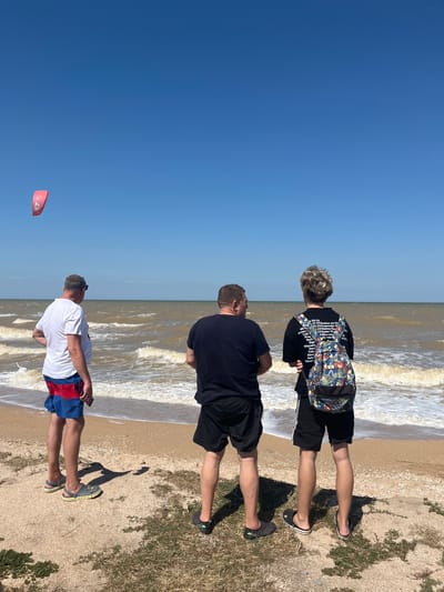 Three individuals are observed from behind, standing on a sandy beach facing a body of water under a clear blue sky. A red kite is visible in the upper left quadrant of the image. The individual on the left wears a white t-shirt and red and blue patterned shorts, with grey and yellow sandals. The middle individual wears a black t-shirt and black shorts, with black slip-on sandals. The individual on the right wears a black t-shirt with a colorful, patterned backpack and black shorts, with black and white slip-on sandals. The sea is moderately choppy with waves breaking on the shore. The location is the beach in Dolzhanskaya, Russia.