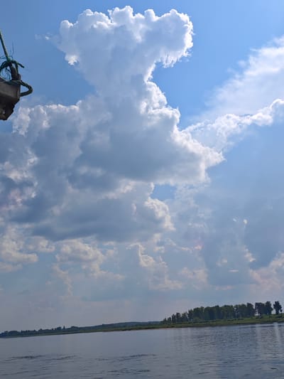 The image captures a wide, calm river in Ust'-Kem', Russia, under a sky filled with dramatic cumulus clouds. The water is a deep blue-gray, reflecting the sky and showing slight ripples on its surface. The far bank of the river is lined with a dense green forest, with a narrow strip of sand visible in places. The sky is a vibrant blue, with large, fluffy white clouds dominating the upper portion of the frame. To the left, a portion of what appears to be a boat or dock structure is visible, with a thick, weathered rope attached. The overall scene conveys a sense of natural beauty and tranquility.