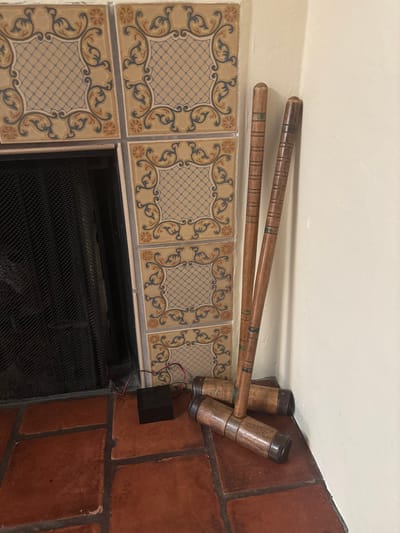 An indoor scene in Monterey, United States of America, depicts a corner of a room featuring a fireplace with a black mesh screen. The fireplace is framed by cream-colored ceramic tiles adorned with blue, gold, and reddish-brown scrollwork and a central blue crosshatch pattern. The floor is covered with terracotta-colored square tiles. Leaning against the light-colored wall to the right of the fireplace are three wooden croquet mallets, each comprising a banded wooden shaft and a cylindrical wooden head with metal end caps. On the terracotta floor, in front of the fireplace opening, a small black rectangular box with connected red and black wires is present. No individuals are visible.