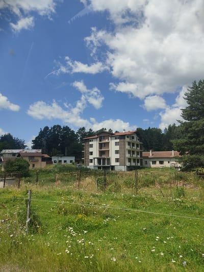 The image captures a scene in the city of Yundola, Bulgaria, on a bright, cloudy day. In the foreground, a wire fence runs horizontally across a field densely populated with tall green grass and small white and yellow wildflowers. The field is bordered by a rough, metal fence in some areas. Behind the field, several buildings are visible. A multi-story, modern building with a white facade and dark-trimmed windows stands prominently, featuring balconies and a reddish-brown tiled roof. To the left of this structure are smaller, older buildings, including one with a similar reddish-brown tiled roof and a pale yellow facade. Further in the background, a dense forest of dark green pine trees forms a natural backdrop, with a tall, narrow structure resembling a water tower or chimney peeking above the treeline in the distance. The sky above is a vibrant blue, punctuated by fluffy white clouds. The overall impression is one of a rural or semi-rural setting with a mix of residential architecture and natural landscape.
