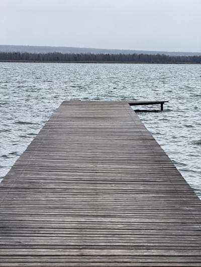 The image depicts an elevated wooden structure or platform, possibly a pier or deck, adjacent to a body of water. The structure is made of parallel wooden planks. A black metal post or railing is located at the edge of the platform where it meets the water. The water's surface has a textured appearance, suggesting movement or waves. In the distance, a strip of land or vegetation is visible, bordered by a hazy sky. Given the context of Kurchatov, Russia, the water body could be part of the Irtysh River or one of the nearby lakes. The scene is still and devoid of any visible people or actions.
