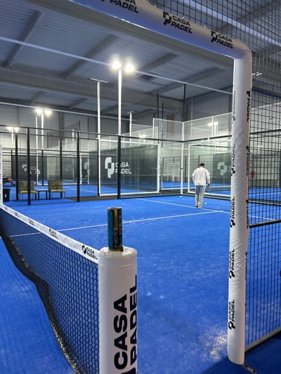 The image captures an indoor padel court with vibrant blue flooring and enclosed by mesh walls. Two padded poles, emblazoned with the "Casa Padel" logo, frame the foreground. A padel racket, partially concealed within the lower pole, hints at gameplay. In the background, a person is visible on the court near the net. The court is well-lit, with overhead lights illuminating the space, contributing to the professional atmosphere of the padel facility.