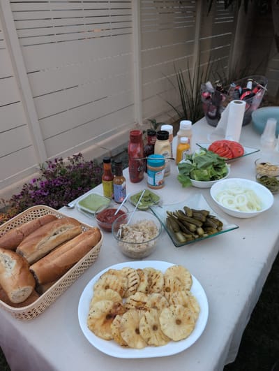 An outdoor setting in Lod, Israel, hosts a buffet table laden with barbecue fixings. A white tablecloth covers the table, which is set against a backdrop of a white slatted fence and lush greenery. The scene captures a spread of food, including bread rolls, grilled pineapple rings, sliced tomatoes, lettuce, onions, pickles, chopped onions, and various condiments such as ketchup, mustard, and what appears to be guacamole. Bottles of beer are visible in a clear ice bucket. The lighting suggests it is late afternoon or early evening. No people are present in the image.
