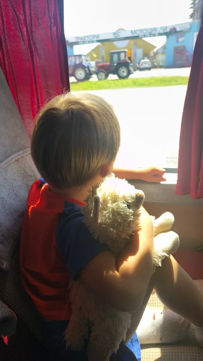 A young child with blond hair, wearing an orange shirt with a blue collar, is holding a fluffy white stuffed animal and looking out a window. The child is seated, and their body is angled to the right. Sunlight streams through the window, illuminating the child's arm and casting shadows. Outside the window, two tractors are visible in front of a building with a sign that reads "TRAKTORI" and "MOTOCIKLI." The setting appears to be a dealership or service center for agricultural and motorcycle equipment in Sokilnyky, Ukraine. The scene is framed by red curtains on the left and right.