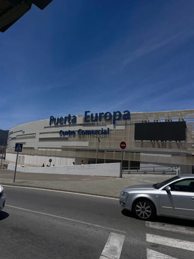 The image shows a street scene in Algeciras, Spain, on a clear, sunny day. The main subject is a modern, white-colored building with large blue lettering that reads "Puerta Europa Centro Comercial." The building appears to be a shopping center. In the foreground, a silver car is parked next to a pedestrian crossing. The car is partially visible, showing its front wheel and part of its body. The sky is a vibrant blue with a few wispy clouds. The overall atmosphere is bright and airy, suggesting it's daytime. There are a few people visible near the building's entrance, but they are too small to discern any details about their activities or emotions. A red circular sign with a horizontal bar through it, commonly indicating a prohibition, is mounted on a pole in front of the building. A pedestrian crossing sign is also visible. The scene conveys a sense of urban activity and commerce.
