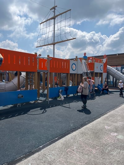 The image shows a playground structure designed to resemble a pirate ship, located in the city of Nagayevo, Russia. The structure is brightly colored with orange and blue elements, and features a mast with netting, slides, ladders, and various platforms. Several people are present on and around the playground. In the foreground, a woman with a pink backpack is standing on the dark grey rubberized surface of the playground. To her right, a woman is sitting on the ground next to a child who is on a ladder. Further to the right, a child in a white shirt and hat is standing. In the background, other children can be seen playing on the pirate ship structure. The sky is partly cloudy with a bright blue sky visible. The ground in the foreground also has chalk drawings on a lighter colored paved surface.