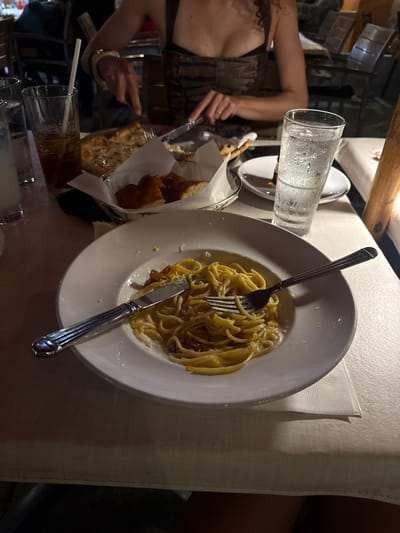The scene shows a table setting with a plate of pasta, likely carbonara, with a fork and knife placed on top. Next to the plate is a basket of bread. There is a glass of water and another drink with a straw. A person is seen in the background, partially visible. The setting appears to be a restaurant or dining establishment. The location is the city of Honolulu, United States of America.