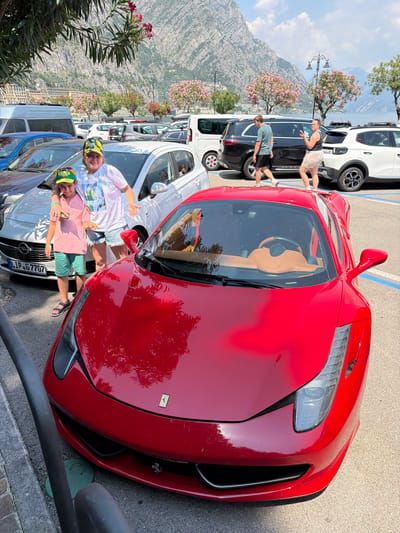 In an outdoor parking lot overlooking a body of water and mountains, two young boys are posing for a photo with a red sports car. The boy in the foreground is wearing a pink shirt, green shorts, and a yellow and green hat. He has his arm around the shoulders of the boy behind him, who is wearing a white t-shirt with a graphic print, denim shorts, and a colorful bandana. Both boys are smiling and appear to be enjoying themselves. They are standing next to a white car, with the red sports car prominently displayed in the center of the image. The parking lot appears to be on a slight incline, with trees and foliage in the background. The sky is a clear blue, suggesting a sunny day. The presence of the red sports car, the style of the cars in the background, and the mountainous landscape with buildings in the distance suggest the location may be a tourist destination in Italy, possibly Limone sul Garda given the provided context. The license plate on the white car reads "LIP G 7707". The scene is brightly lit, indicating it is daytime.