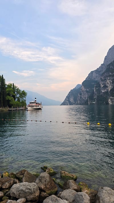 This image depicts a serene waterscape at Riva del Garda, Italy, likely during late afternoon or early evening due to the soft lighting and pastel hues in the sky. The scene is dominated by a calm, clear lake, reflecting the soft light. In the distance, imposing, tree-covered mountains rise dramatically, their slopes disappearing into a hazy sky.

A prominent feature is a white and red passenger boat docked at a wooden pier, surrounded by lush green trees. The boat appears to be waiting for passengers or has just arrived. A line of red and white buoys stretches across the water, marking a swimming area or a navigational channel. The foreground is characterized by a rocky shoreline, with large, gray stones, some covered in green moss, meeting the water's edge. The water is shallow near the shore, allowing a glimpse of the submerged rocks.

The sky is a soft blue, streaked with wispy, white clouds, some of which are tinged with the warm colors of sunset. The overall mood is peaceful and tranquil, evoking a sense of calm and natural beauty. No visible text is present in the image.