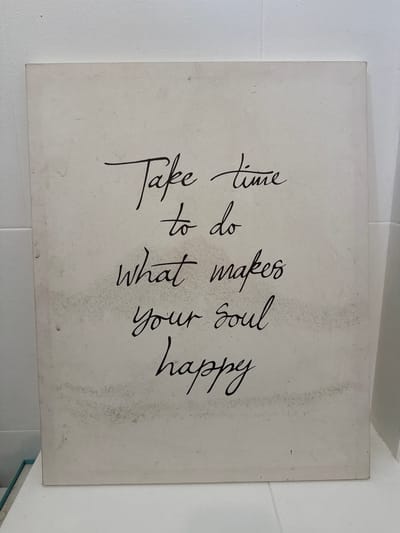 A framed canvas sign with a quote written in black cursive handwriting is displayed against a white tiled wall. The quote reads, "Take time to do what makes your soul happy." The canvas is off-white and shows some light staining or discoloration in patches, particularly towards the bottom left and right edges. The text is centered on the canvas. The scene appears to be indoors, possibly in a home or a shop. There are no people or animals visible in the image. The lighting is neutral, suggesting an ambient indoor environment. The overall impression is of a decorative item providing an inspirational message. The location of Tha Chana, Thailand, is not discernible from the image itself; this information is external to the visual content.