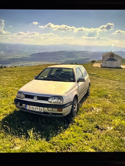 A silver Volkswagen Golf Mk3 is parked on a grassy hill overlooking a valley. To the right of the car, a small white building with a blue dome and a cross on top sits on the hill. The sky is blue with scattered white clouds, and the sun is casting shadows. The license plate of the car reads "CB 8649 BA". The car is facing slightly to the right, and the building is in the background. The overall scene is serene and picturesque, likely in a rural area near Dragoman, Bulgaria, given the distinctive license plate format common in Bulgaria.