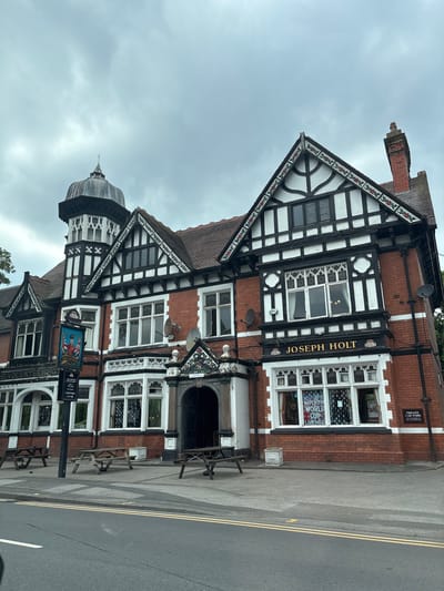 This is a photograph of a traditional Tudor-style public house, likely a pub, with a distinctive dark timber framing and white render against a red brick facade. The building features large, ornate windows with leaded glass and a prominent gabled section. A small dome or cupola is visible on the left side of the building. The building appears to be situated on a street with a road and pavement.
Visible text on the building includes "JOSEPH HOLT" and "THE VOLUNTEER HOT". There are also signs indicating "PRIVATE CAR PARK" and "CAR PARK". Other smaller signs mention "HOLT" and depict a soldier.
The time of day appears to be daytime, with an overcast, cloudy sky suggesting possibly dull or inclement weather. The scene appears quiet, with no people visible in the immediate vicinity, though there are several outdoor picnic-style tables on the pavement. The presence of outdoor seating suggests the pub may be open for business, but the overall atmosphere is somewhat subdued. The location is indicated as Trafford, United Kingdom, which aligns with the style of the architecture commonly found in that region.