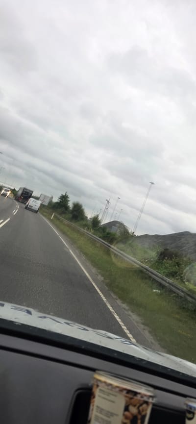The image depicts a roadside view from inside a vehicle, likely traveling on a highway in Esbjerg, Denmark, given the presence of wind turbines, a common sight in the region. The sky is overcast with heavy, gray clouds. The road ahead is visible, with multiple lanes and white lane markings. Several vehicles are on the road, including a white van and a dark truck with "FAIRTRANS" written on its side.
On the right side of the road, there is a grassy embankment with a metal guardrail. Beyond the embankment, a large pile of dark material, possibly coal or another industrial product, is visible. Several wind turbines are erected in the distance, some appearing operational and others as static structures. Trees and other vegetation are scattered along the roadside.
The foreground of the image shows the interior of the vehicle, including the dashboard and part of a windshield, which has some streaks or reflections. A cylindrical container, possibly for snacks, with a label featuring images of nuts, is placed on the dashboard. The overall scene suggests a cloudy day, with traffic on a highway near an industrial or energy generation area.