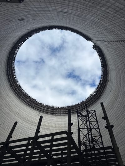 This is an interior view looking up at the cooling tower of the Chernobyl Nuclear Power Plant, located in Pripyat, Ukraine. The massive, conical structure is made of concrete and is partially constructed. The opening at the top reveals a cloudy sky with patches of blue. Inside the tower, there is scaffolding and industrial metal framework, suggesting ongoing or halted construction. The scale of the structure is emphasized by the low angle and the vastness of the concrete walls curving upwards. There are no people visible in the image. The scene is desolate and industrial, with a sense of abandonment or incomplete development.