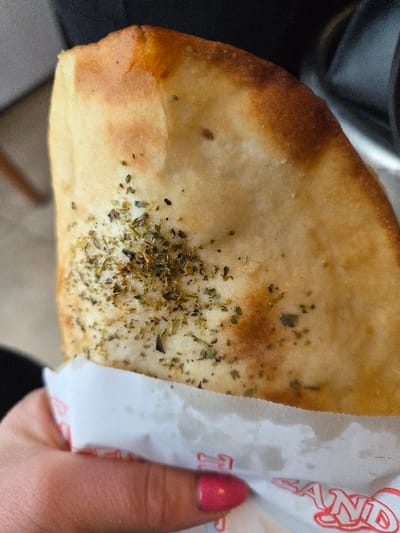 A human hand, with fuchsia-painted fingernails, holds a triangular portion of a baked dough product, likely a flatbread or calzone, in Gevgelija, North Macedonia. The product's golden-brown crust shows uneven baking, with a lighter underside visible, and its surface is liberally dusted with dried green herbs. A white paper wrapper, featuring red text including the partial word "SAND", encases the lower section of the food item. The background is blurred, indicating an indoor setting with indistinct dark and light elements.