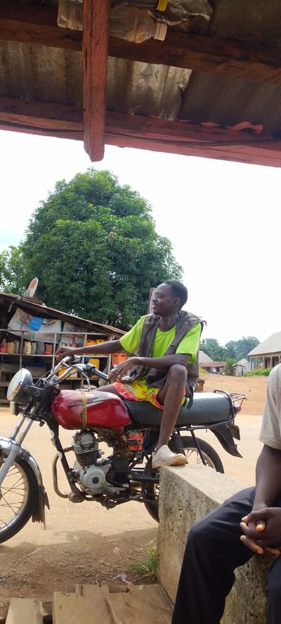 A young man with dark skin, wearing a bright lime green t-shirt and a dark vest, is sitting on a red motorcycle. He has on yellow shorts with red and white patterns and white shoes. He is looking to his right, with a slight smile. The motorcycle is parked in front of a low concrete barrier. Behind the motorcycle, shelves are stocked with various colorful plastic containers, likely fuel or oil cans, suggesting a roadside stand or small shop. A large, leafy green tree is visible in the background. The sky is overcast. The setting appears to be an outdoor market or roadside area in Baissa, Nigeria.