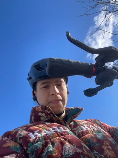 A low-angle photograph taken in Altafulla, Spain, depicts a male individual and bicycle components against a clear blue sky. The male, positioned in the lower frame, wears a dark blue bicycle helmet and a burgundy corduroy puffer jacket patterned with green, gold, and white floral motifs. The word "KARL" is embroidered in white on the jacket's chest. An earbud is visible in his left ear. He looks directly at the camera with his tongue slightly extended. To the right, black bicycle handlebar components are visible, including a rubber grip, a black brake lever with visible damage near its tip, and a gear shifter. A red ring accents the base of the handlebar grip. The background consists of a bright blue sky with scattered white clouds and bare tree branches.