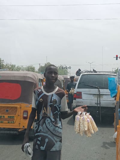 A young male stands centered on a paved road in Hotoro, Nigeria, facing the viewer. He wears a short-sleeved, v-neck t-shirt with brown and light blue geometric patterns, featuring text that includes "SAINT-GERMAIN" and "PSG". In his left hand (viewer's right), he holds a cluster of approximately 10-12 small, clear plastic bags containing a light-colored, granular snack, consistent with popcorn. In his right hand (viewer's left), he holds a small, white plastic bag containing an unidentifiable object.
Behind the primary subject, a row of yellow and brown auto-rickshaws are visible on the left side of the frame, appearing to be stationary in traffic. To the right of the primary subject, a light-colored SUV, with "CITROËN" visible on its rear, is also stationary. Another male individual, dressed in dark clothing, is positioned between the primary subject and the SUV, also holding bags of merchandise. A third individual is visible further back on the roof rack of the SUV. The background consists of an overcast sky, distant trees, utility poles, and traffic lights displaying a red signal on the far right. The scene depicts a bustling urban street environment during daylight hours.