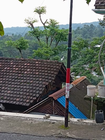 The image depicts an outdoor scene in Bulukandang, Indonesia, presenting an elevated view over a residential area backed by forested hills. In the immediate foreground, a dark asphalt surface occupies the lower left corner, transitioning to a light grey concrete ledge that runs horizontally across the lower frame. A vertical, dark-colored pole, featuring a red and white banded section near its base, stands prominently on this ledge, extending into the upper midground. Green string is wrapped around the pole mid-section, and moss is present at its base. A light-colored rock fragment rests on the concrete ledge to the left of the pole. To the right, on a lower concrete platform, a dark plastic jerry can is situated, with two white plastic buckets on a higher, moss-covered platform further right.
Below the concrete ledge, occupying the midground, are several building structures. The most prominent is a roof composed of dark brown, overlapping clay tiles, partially covered with moss and dirt. This tiled roof slopes downwards from left to right. Beneath and to the right of this tiled roof, sections of other roofs and building sides are visible, including a rust-colored corrugated metal roof and a distinct bright blue corrugated metal roof section. Exposed wooden paneling is also apparent on the side of one of these lower structures.
The background is dominated by dense green foliage and numerous trees, obscuring any distant buildings. A range of undulating hills, entirely covered in dark green forest, extends across the horizon under an overcast or hazy sky. No individuals or animals are present in the visible scene.