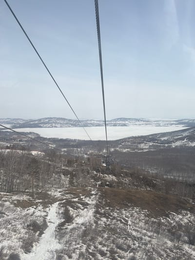 The image displays a panoramic view from a ski lift cabin traversing a winter landscape at a ski resort in Zelenaya Polyana, Russia. Two prominent steel cables of the ski lift system extend from the upper left and right, converging towards the lower center. The cable on the right is thicker and exhibits a ribbed texture.
Below, a descending mountain slope is visible, characterized by patchy snow cover interspersed with areas of exposed brown, dry vegetation. A narrow, winding track in the snow is discernible on the lower left portion of the slope. In the midground, a mixed forest composed predominantly of bare deciduous trees with lighter bark and some darker evergreen trees is present. Several grey-colored ski lift gondola cabins are suspended from the cables, moving away from the viewer down the slope. Structures resembling resort buildings and additional ski lift support infrastructure are partially visible among the trees on the left side of the midground.
The background features a vast, frozen body of water, consistent with Lake Bannoe, completely covered in white snow or ice, stretching across the horizon. Beyond the frozen lake, a range of snow-dusted hills or mountains forms the distant backdrop under a pale blue, slightly hazy sky. No individuals are discernibly present or interacting in the frame.