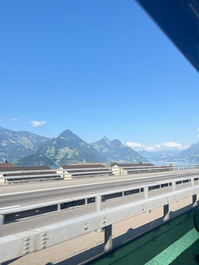 The image presents a panoramic daytime view from a moving vehicle on a multi-lane highway or bridge in Buochs, Switzerland. In the immediate foreground, the lower right corner shows a dark, possibly tinted window frame of the vehicle. A light-gray metal guardrail with exposed bolts runs horizontally across the mid-lower section, casting distinct shadows onto the concrete road surface, which features two visible lanes marked with white lines. Beyond the highway and guardrail, a row of light-yellow or beige single-story buildings with brown sloped roofs are visible. These structures are situated adjacent to a large body of calm, blue water, identified as Lake Lucerne. The background is dominated by a clear, bright blue sky with a few small, scattered white clouds. A range of prominent mountains, characteristic of the Swiss Alps, rises from the lake's edge, exhibiting lush green vegetation on their slopes and some visible rock formations. The scene is brightly illuminated by direct sunlight. No individuals are present.