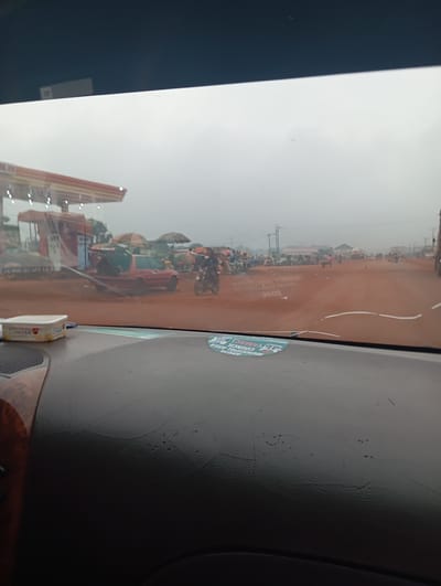 The image, taken from the interior of a vehicle, shows a dusty, overcast scene in Imilike, Nigeria. A gas station with an orange and white awning is visible on the left. In front of it, a red car with its trunk open is parked on a dirt road. Several people and motorcycles are present near the gas station and further down the road. The road appears to be a busy thoroughfare with market stalls and other vehicles. The windshield of the car has a crack and several stickers are visible on the dashboard, one with the text "ABUJA MUNICIPAL COUNCIL".
