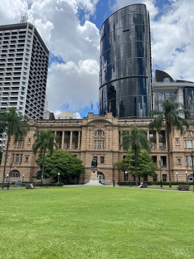 This image captures a vibrant urban scene in Brisbane City, Australia, showcasing a blend of architectural styles and natural elements. The scene depicts a large, well-maintained green lawn on the left, flanked by prominent buildings on the right and in the background.

On the right side of the frame stands a grand, ornate historic building constructed from light-colored stone. It features classical elements such as columns, arched windows, and intricate decorative details, suggesting it might be a government or institutional building. A dark statue on a light pedestal is visible at its base, and benches and streetlights line the path in front of it. Behind and slightly to the left of this historic structure is a modern, sleek skyscraper with a curved facade of dark, reflective glass. A distinctive stylized star or burst logo is visible near the top left section of this contemporary building. Further in the background, on the far left, another tall, rectangular modern building with a grid-like pattern of windows and concrete frames contributes to the city skyline.

The expansive green lawn on the left side of the image is immaculate, providing a strong contrast to the urban structures. Several palm trees and other leafy green trees are interspersed among the buildings and along the edges of the lawn. One person is visible, seated on a dark bench on the path between the historic building and the lawn, seemingly relaxed and enjoying the surroundings.

The image portrays a typical daytime scene, with bright sunlight creating clear shadows, suggesting late morning or early afternoon. The sky is partly cloudy, featuring patches of blue interspersed with white clouds, indicating fair weather. The unique juxtaposition of the historic and modern architecture, particularly the ornate building and the curved glass skyscraper, along with the extensive green space, serves as a clear location cue for Brisbane City. The overall atmosphere conveys a sense of urban development and historical preservation coexisting, with opportunities for leisure in the public green areas. No explicit readable text is visible, apart from the stylized star logo on the modern skyscraper.