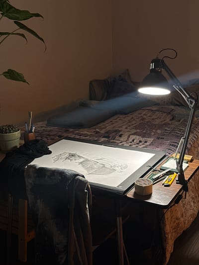 The image depicts a drawing setup indoors. A black articulated lamp is positioned above a drawing board, illuminating a work-in-progress drawing of a figure on white paper. The drawing board rests on what appears to be a bed, partially covered by patterned bedding. Various drawing tools and supplies, including pencils, a ruler, and masking tape, are placed on the board alongside the drawing. A cactus in a pale green pot is visible in the lower foreground, along with plant leaves. The room is dimly lit, with the primary light source being the lamp. The location is not identifiable as Montreuil, France, based on the visible contents of the image.