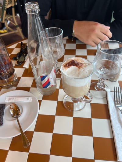 The image presents a tabletop scene at a cafe in Deauville, France. The table surface features a distinct brown and white checkered pattern. Arranged on the table are multiple items. On the left, a partially visible clear glass bottle containing a dark liquid with ice is present, alongside a pair of folded black sunglasses. Adjacent to these are a small white ceramic plate holding a golden spoon, a white sugar packet labeled "SUCRE," and a dark wrapper. In the center, an empty tall clear glass bottle with a red, white, and blue striped label stands upright. To its right, an empty stemmed glass is visible. A stemmed glass mug contains a layered beverage, appearing as light brown liquid topped with a thick layer of white foam and sprinkled with dark powder. On the far right, another stemmed glass holds a small volume of clear liquid, positioned next to a white folded napkin and a silver fork. In the upper right background, a person's forearms and hands are visible, resting on the table. The person wears a long-sleeved black garment and a light-colored bracelet on the left wrist. Blurred background elements suggest an outdoor cafe setting with additional seating.
