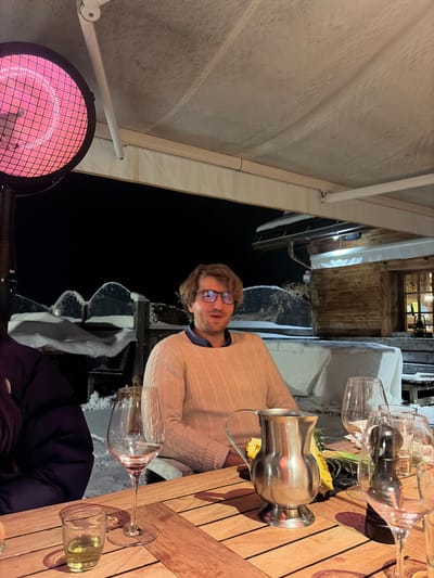 The image depicts an outdoor dining scene at night in La Clusaz, France. The foreground shows a wooden slatted table with several items: an empty wine glass to the left, a small clear shot glass containing a yellow liquid, a silver-colored metal pitcher, and another wine glass along with a black pepper mill on the right. A male individual with light brown hair and eyeglasses, wearing a light beige cable-knit sweater over a collared shirt, is seated at the table, looking forward with a slightly open mouth. To his left, the shoulder and arm of another person, clad in a dark purple or black puffer jacket, are partially visible. A round, red-glowing electric patio heater stands prominently on the far left. Above the scene, a light-colored retractable awning or canopy covers the dining area. The background reveals a snow-covered landscape under a dark night sky. Snow-laden structures, possibly benches or low walls, are visible behind the man. To the far right in the background, a rustic wooden chalet-style building with a lit window can be seen. The overall environment suggests a cold, winter setting, with artificial heating provided for comfort.