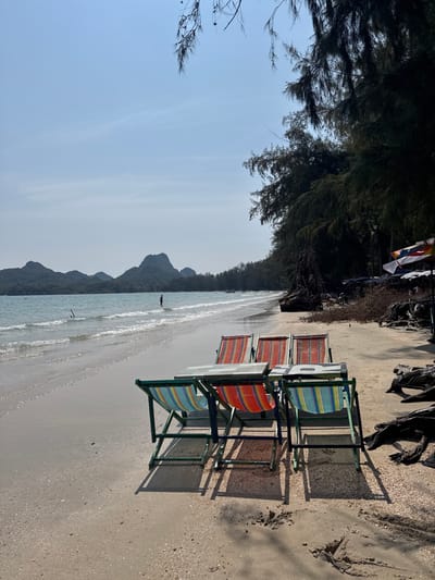 A beach scene features four empty lounge chairs, each with a colorful striped seat and a green frame. They sit in the sand, casting long shadows. The ocean stretches into the distance, framed by mountains and trees. The sky is blue and mostly clear. The chairs face towards the water, creating a tranquil and inviting setting.