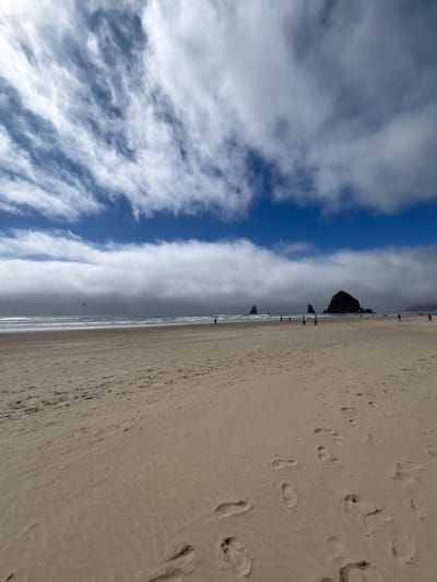 A vast and serene beach scene at Cannon Beach, United States, unfolds under a dynamic sky. The image, best viewed with a 90-degree clockwise rotation, prominently features a wide expanse of light-colored, sandy beach in the foreground and mid-ground. The sand is marked with numerous footprints, indicating recent human activity along the shoreline.
In the mid-distance, the gentle waves of the Pacific Ocean meet the sand, creating a crisp horizon line. Scattered along this waterline are several small figures of people, seen walking leisurely, suggesting a calm and recreational atmosphere. The iconic Haystack Rock stands prominently further down the coastline, accompanied by smaller sea stacks, serving as a distinct geographical identifier for Cannon Beach.
The upper portion of the image is dominated by the sky, which exhibits a striking contrast. A horizontal band of clear, vibrant blue sky runs across the upper middle of the frame. This azure strip is flanked by various cloud formations: above it, delicate, wispy white clouds stretch across the expanse, while below it, denser, whiter, and light grey clouds gather.
The lighting suggests it is daytime, likely late morning or early afternoon, with ample natural light illuminating the scene. There is no visible text in the image. The overall impression is one of peaceful coastal enjoyment amidst varied weather conditions.
