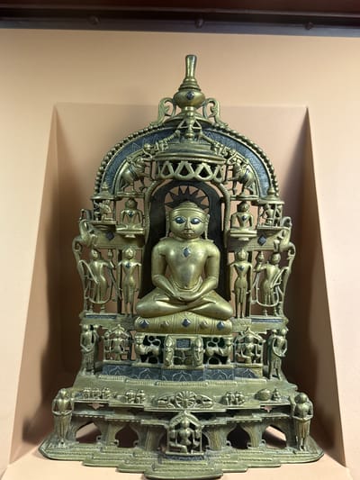 The image depicts a bronze sculpture, possibly of a Jain Tirthankara, mounted on a wall in what appears to be a museum or gallery setting. The sculpture is highly detailed, with intricate carvings and multiple figures surrounding the central figure, which is seated in a meditative pose. The location is specified as Lalitpur, Nepal, which is known for its rich artistic heritage and metalwork. The wall behind the sculpture is a neutral beige color, and the lighting is diffused, suggesting an indoor environment designed to showcase art.