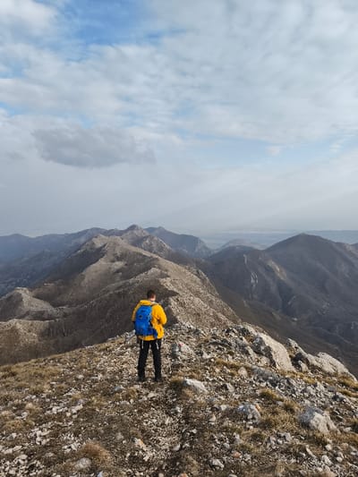 The image displays a solitary male figure standing on a rocky mountain ridge, facing away from the viewer, in the vicinity of Mali Mikulići, Montenegro. The individual wears a bright yellow jacket, black trousers, and a blue backpack, and appears to be utilizing trekking poles. He is positioned on a rugged, uneven surface composed of grey and white rocks and sparse, dry brown grass. A red and white trail marker is visible on a rock to the right of the figure. The background reveals a vast panorama of undulating mountain ridges and peaks, characterized by dark, sparse vegetation and exposed rock, receding into an atmospheric haze. The sky transitions from a partly cloudy blue with scattered white cumulus clouds in the upper portion to a hazy, lighter tone near the distant horizon, which appears to feature a flatter landmass or water body. The overall scene is illuminated by diffuse daylight.