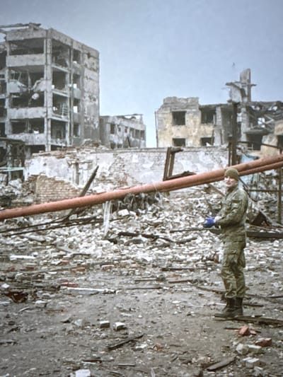 A person wearing a camouflage uniform and a beanie stands amidst the debris of destroyed buildings. The buildings appear to be multi-story structures with damaged facades, broken windows, and exposed interiors. The ground is covered in rubble, including bricks, concrete, and wooden debris. A large orange pipe runs horizontally across the scene, supported by debris. The sky is overcast. The location in the city of Pori, Finland is not discernible from the image.