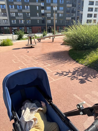 This media file captures a view from a stroller, looking down at a baby and then outwards to a modern apartment building and a playground. The baby, swaddled in a light-colored blanket and wearing a yellow outfit, is the primary subject within the stroller. The stroller itself is dark blue.
The scene is set outdoors on a sunny day, indicated by the distinct shadows of trees cast on the reddish-brown pavement. The playground area features chalk drawings of hopscotch squares. In the background, a multi-story apartment building with a contemporary architectural design dominates the view. The building has a facade with alternating colors of gray, white, and dark panels, featuring numerous windows. Greenery, in the form of bushes and trees, is visible around the building and on the playground.
The lighting suggests it is daytime, likely morning or afternoon, given the strong shadows. The overall mood appears peaceful and domestic, with the focus on the child and the surrounding urban environment. There is a sign visible on the apartment building with Cyrillic text that appears to read "СТАРТ," which translates to "START." The location context provided is Patrusheva, Russia.