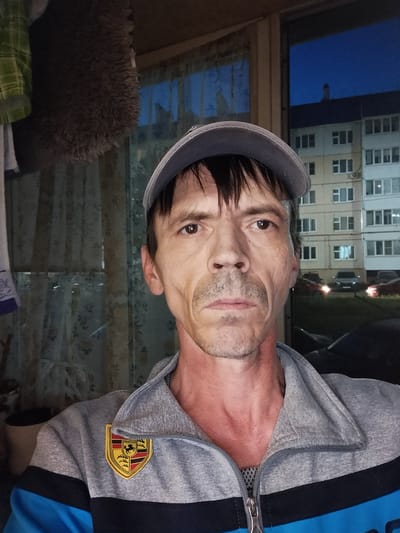 This is a selfie taken by a man outdoors at dusk in Zyuzino, Russia. The man is wearing a grey baseball cap and a grey and blue zippered jacket with a Porsche crest on the left chest. He has a thin build and fair skin, with visible lines on his face and a reddish hue on his neck. His expression is neutral.
In the background, we see a multi-story apartment building with many windows, typical of Russian urban landscapes. Several cars are parked in front of the building, and the sky is a deep blue, indicating it is evening or twilight. The setting appears to be a residential area. There is no explicit indication of any activity or event, other than the man taking a photograph of himself.
