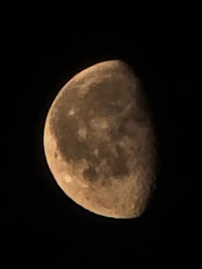 The image shows a close-up view of the moon against a black sky. The moon is partially illuminated, with roughly half of its surface visible. The visible surface of the moon displays a textured appearance with darker and lighter patches, indicative of craters and lunar terrain. The lighting suggests the moon is in a waxing or waning phase, possibly a gibbous or nearly full moon. The color of the moon appears to be a warm, desaturated yellow-orange, possibly due to atmospheric conditions or photographic processing. There are no identifiable landmarks, buildings, or people in the image, as the focus is solely on the celestial body. The background is completely dark, emphasizing the moon as the sole subject. The image does not contain any information or context related to the city of Икское Устье, Russia.