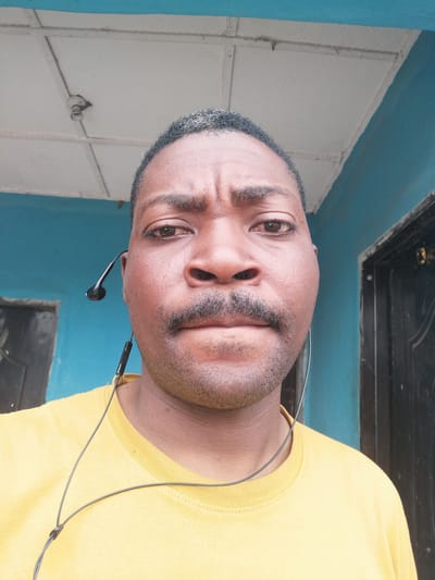 In Ajasse Ipo, Nigeria, a selfie is taken from a medium shot of a Black man. He is wearing a yellow t-shirt and black earbuds. The background consists of a blue wall and a white ceiling with a light fixture. The man has a mustache and short hair. The lighting is natural and appears to be daylight.