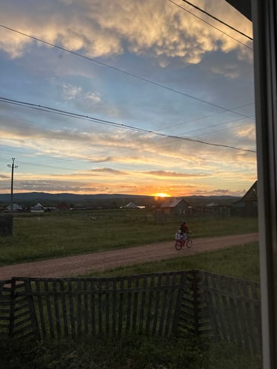 This media file shows an outdoor scene in Urazovo, Russia. The image is oriented vertically, with the sky and power lines on the left and a dirt road, grassy field, and fence on the right. Two people are riding bicycles on the dirt road. The sky is filled with dramatic clouds, illuminated by a sunset. The sun is setting behind a distant mountain range, casting a warm glow across the sky. The village houses are visible in the background, nestled amongst the hills. The overall mood is peaceful and serene, with a sense of quiet beauty. The time of day is sunset. The weather appears clear and pleasant. There is no visible text in the image.