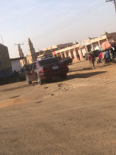 The scene depicts an active, sunlit street in Tukur Tukur, Nigeria. In the foreground, a dark red (maroon) Mercedes-Benz C230 sedan, license plate "ABC 195AV," is angled on a weathered, light brown road surface, showing visible potholes and a textured concrete/metal grate to its right. To the left, a blue truck with a tan canvas cover is parked. Two prominent, light-colored minaret-like structures rise in the mid-distance on the left, accompanied by utility poles and overhead lines.
The background features a row of single-story commercial buildings with peach/pink facades and some blue trim, lining the street. A sign bearing "UCHE" is visible on one building. Further right, an open-air market area is present, indicated by an orange parasol sheltering vendors and goods.
Several people are visible: one individual walks towards the right foreground with a walking stick, casting a distinct shadow. Others are gathered around the market stall under the parasol. A person in light-colored attire is visible near a partially obscured white pickup truck (possibly a Ford) in the background on the right. The sky is a pale blue, suggesting clear conditions, with shadows indicating either mid-morning or late afternoon.