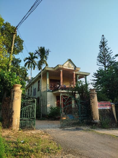 The image depicts a two-story, pale yellow residence situated on a dirt road in Đăk Xú, Vietnam, under a clear blue sky. The building features a prominent second-floor balcony supported by two reddish-brown fluted columns, topped by an ornate pediment with light-colored relief sculpture. Several dark-tiled solar panels are visible on the lower roof section of the balcony. The ground floor entrance has dark wooden doors and a small overhang.
Access to the property is via a set of weathered green metal gates, partially open, flanked by two substantial, weathered light-brown concrete pillars. A rectangular dark panel with a red sign, partially legible as "HỘP PHÁT CUỘC..." in Vietnamese, is affixed to a dark, moss-covered wall section adjacent to the right pillar.
Lush green vegetation surrounds the property, including large leafy trees on the left, a wooden utility pole with multiple overhead power lines, two tall areca palm trees positioned between the gate and the house, and a tall, conical evergreen tree on the right side of the building. The foreground consists of a dirt and gravel road with grassy verges. No individuals are present, and the scene is static under natural daylight.