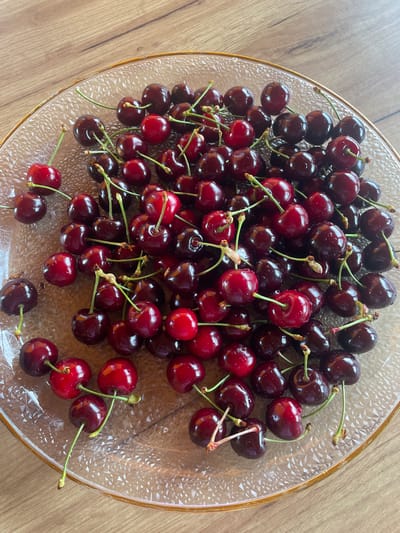 A transparent, textured glass plate with a gold-colored rim contains a substantial quantity of fresh cherries. The cherries, predominantly dark red to deep maroon, exhibit varying shades with some brighter red examples interspersed. Numerous small water droplets are visible on the skin of many cherries, indicating recent washing or freshness. Most cherries retain intact green stems, some individually attached, others in small clusters. The plate, filled mostly with the cherries, rests on a light to medium brown wooden surface, displaying discernible wood grain patterns. The illumination suggests natural light, creating soft reflections on the fruit and glass.
