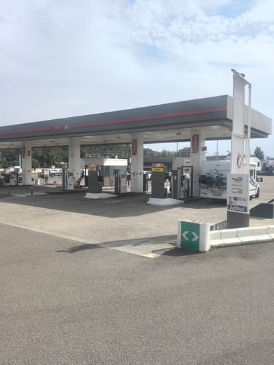 A view of a TotalEnergies gas station on a cloudy day in Solaize, France. The gas station features multiple fuel pumps under a covered canopy. A white camper van with a bicycle rack carrying two bicycles is parked at one of the pumps. Signage indicates services such as "Diesel" and "Air". The ground is paved asphalt. In the foreground, a white concrete barrier with green directional arrows is visible. The background shows trees and a hillside.