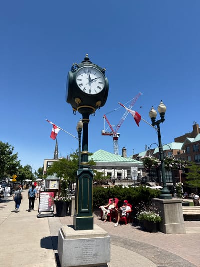 The image captures a bright, sunny day in downtown Oakville, Canada, likely during the summer. A prominent, dark green and gold ornate clock stands in the foreground, displaying the time as approximately 2:00 PM. The clock is situated on a stone pedestal with an inscription that reads "CLOCK SPONSORS" followed by a list of local businesses and organizations.

To the left of the clock, a paved walkway is visible, with several pedestrians walking along it. Two women, one in a grey shirt and jeans, the other in a blue shirt and black pants, are walking away from the camera. Further down the walkway, a sign titled "What's On" is displayed, detailing upcoming events.

In the background, several buildings with green roofs and brick facades line the street. Two Canadian flags are prominently displayed, one on a flagpole near the clock and another on a construction crane in the distance. The crane suggests ongoing construction in the area.

Several people are seated on red Adirondack chairs in a landscaped area to the right of the clock, enjoying the weather. A lamppost adorned with hanging flower baskets stands next to the clock. The sky is clear blue, indicating good weather conditions.