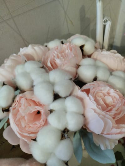 A close-up shot captures a floral arrangement of light pink peonies and fluffy cotton bolls. The peonies, with their many delicate petals, exhibit soft shades of pink, some appearing lighter than others. Interspersed among the flowers are the cotton bolls, their white, airy texture contrasting with the floral elements. Some of the cotton bolls show dark brown centers. A few muted green leaves are also visible, adding a subtle touch of color to the arrangement. The background is a textured, light beige wall. To the right of the frame, a white object with a silver metallic detail is partially visible, possibly a part of a larger object or decoration. The lighting is soft, suggesting an indoor setting, possibly with ambient light. No people are visible in the frame. The scene evokes a sense of delicate beauty and natural textures. The location context is Щучье, Russia, but there are no specific visual cues in the image to confirm this. There is no visible text in the image.