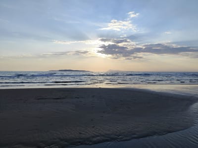 The image shows a beach scene at Agios Stefanos, Greece. The sky is blue with scattered clouds, and the sun is partially obscured by the clouds, creating rays of light. Below the sky is the sea with gentle waves. Two small islands are visible in the distance. The foreground shows the sandy beach, with wet sand in the immediate foreground displaying ripple patterns. The overall lighting indicates either sunrise or sunset. There are no people or animals visible in the image.